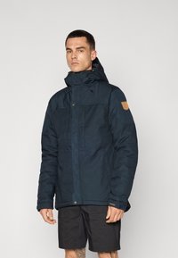 Dark blue insulated jacket with a high collar and adjustable hood. Textured fabric, zippered front, and logo patch on the sleeve.