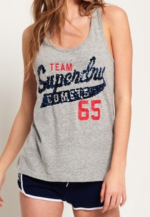 Kobieta w szarej koszulce na ramiączkach z napisem "Team Superdry 65" i niebieskich szortach z białą lamówką, stoi na jednolitym tle.