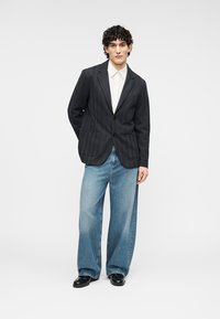 Blazer noir texturé avec deux poches avant, associé à un jean large bleu clair. Le mannequin est debout sur un fond blanc uni.