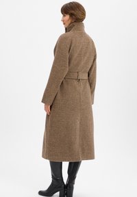 Frau in einem langen braunen Hahnentrittmuster-Mantel mit hohem Kragen und schwarz-gestiefelten knielangen Stiefeln, seitlich auf einem weißen Hintergrund stehend.