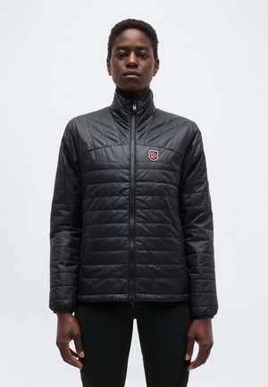 Άτομο που φοράει μαύρο πουπουλένιο μπουφάν με φερμουάρ μπροστά και κόκκινο λογότυπο αλεπούς στο στήθος, στέκεται μπροστά σε απλό ανοιχτό φόντο.