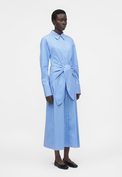 Γυναίκα που φοράει μπλε και λευκό ριγέ φόρεμα με κουμπιά, μακρυμάνικο και με έναν μεγάλο δεμένο κόμπο στη μέση, καθώς και μαύρα φλατ παπούτσια.