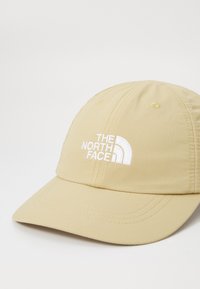 Beige sapka tartós anyagból, ívelt karimával készült. Elöl fehér hímzett logóval és varrási díszítéssel rendelkező.