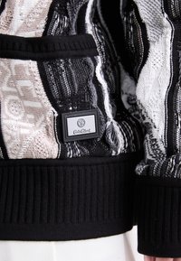 Black and white patterned knit jacket with a ribbed black hem, featuring a rectangular label on the side with the brand name.