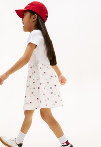 Παιδί με λευκό τοπ και φόρεμα με σχέδιο από κόκκινα λουλούδια. Φορά κόκκινο καπέλο, λευκές κάλτσες και παπούτσια με μαύρα και μπεζ στοιχεία.