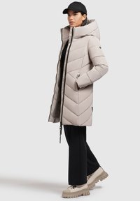 Beige gewatteerde parka met een hoge kraag, chevronpatroon, zijzakken en ritsluiting, gecombineerd met zwarte broek en chunky beige sneakers.