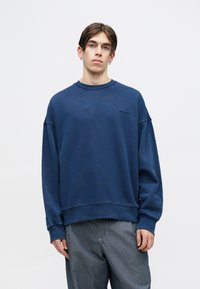 Blauer Sweatshirt mit Rundhals, langen Ärmeln und gerippten Bündchen. Mit dezenter Markenlogo auf der Brust. Kombiniert mit grauen Hosen.