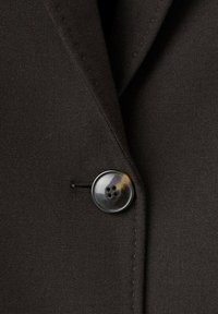 Ciemnobrązowa tkanina kurtki zbliżenie na okrągły czarny guzik z czterema dziurkami oraz subtelnymi różnicami w odcieniu i fakturze.