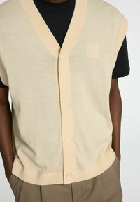 Bež pletena brezrokavnik z globokim V-izrezom, kratkimi rokavi in gumbnim zapiranjem, v kombinaciji s črnim T-shirtom spodaj. Nežen tekstura.