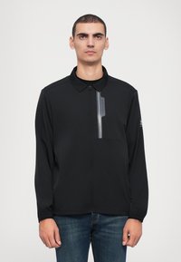 Casaco leve preto com fecho à frente, colarinho, bolso no peito e detalhe refletor. Textura suave e design aerodinâmico.