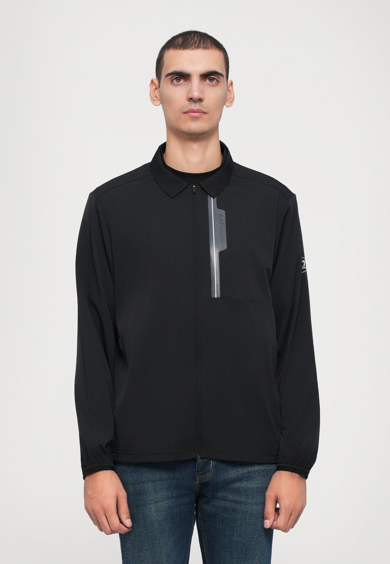 Casaco leve preto com fecho à frente, colarinho, bolso no peito e detalhe refletor. Textura suave e design aerodinâmico.
