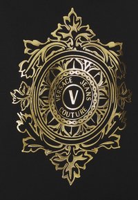 Emblema in foglia d'oro con design ornamentale con foglie, cerchio centrale con il testo "VERSACE JEANS COUTURE" su sfondo di tessuto nero.