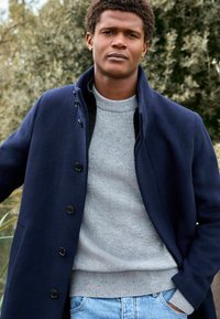 Cappotto in lana blu navy con bottoni neri, indossato sopra un maglione grigio chiaro. Il cappotto presenta un colletto alto e una vestibilità rilassata, abbinato a jeans di denim.