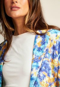 Giacca di tessuto multicolore con motivi astratti blu, gialli e arancioni, indossata sopra una maglietta bianca con una consistenza morbida.