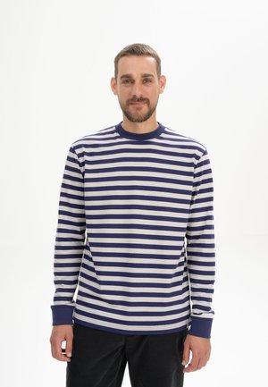 Man met kort haar en baard, gekleed in een marineblauw-wit horizontaal gestreept shirt met lange mouwen en zwarte broek, staand tegen een effen witte achtergrond.