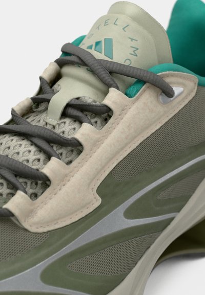 Acercamiento a una zapatilla deportiva verde y beige con tejido de malla, cordones negros y forro interior en color aguamarina, mostrando detalles de costura y acentos de diseño.