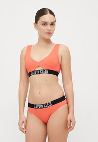 Oranžinė juostuota bikini viršutinė dalis su V formos kaklu, su juodu elastingu juosmeniu su 'CALVIN KLEIN' logotipu; suderinamas bikini apatinias dalis su panašiu dizainu.