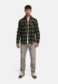 Chemise en flanelle à carreaux verts et noirs avec deux poches poitrine, portée sur un t-shirt blanc, associée à un pantalon gris clair et des baskets vertes.