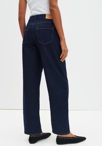 Jeans in denim a vita alta con gamba larga in blu scuro, con una toppa in pelle color cuoio sul retro e cuciture arancioni a contrasto.