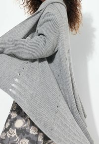 Šedý žebrovaný cardigan s kapucí, volným střihem a spadlými švy na ramenou. Texturovaný vzor pleteniny s otevřenými detaily stehu.