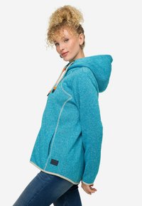 Felpa in pile di colore teal con cappuccio regolabile, dettagli a contrasto di colore chiaro, fessure laterali e patch con logo. Tessuto morbido e vestibilità casual.