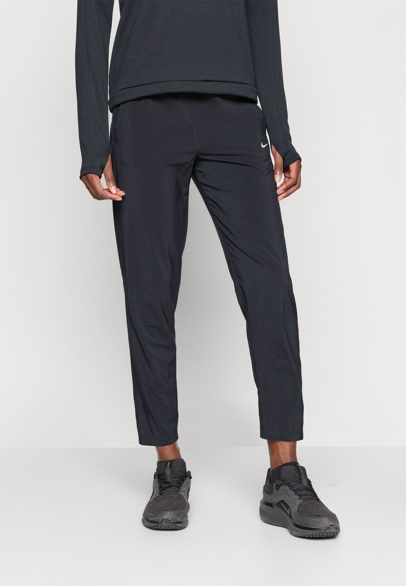 Personne portant un pantalon de sport Nike noir et des baskets noires, debout devant un fond clair uni.