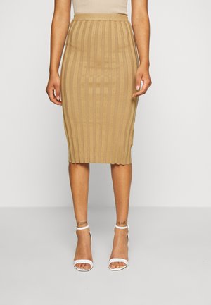 Jupe crayon en tricot couleur beige clair avec une texture en côtes verticales ; longueur mi-mollet, taille élastique ; associée à des talons blancs à lanières.
