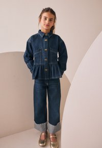 Fiatal lány áll zsebre dugott kézzel, sötét farmerdzsekit arany gombokkal, felhajtott farmernadrágot és arany cipőt viselve, semleges háttér előtt.