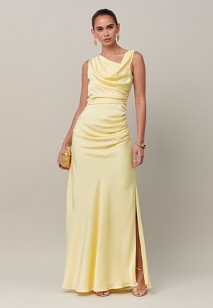 Vrouw die een gele mouwloze satijnen galajurk met split aan de zijkant draagt, een gouden clutch vasthoudt, geaccentueerd met gouden oorbellen en armband.
