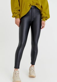 Pantalones skinny de imitación de cuero negros con una cintura alta y textura brillante, combinados con un sudadera holgada verde oliva y zapatillas beige.
