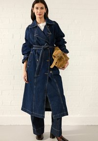 Denim trench kaput s dvostrukim kopčanjem, širokim ovratnikom, pojasom u struku, bočnim džepovima i kontrastnim šavovima; u kombinaciji s odgovarajućim trapericama.