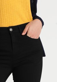 Jeans negros con un diseño ajustado, que cuentan con un bolsillo frontal y un cierre de botón metálico. Combinados con una blusa amarilla de canalé texturizada.