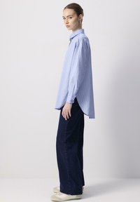 Giovane donna in piedi di lato, con indosso una camicia larga a strisce blu e bianche, pantaloni neri a gamba larga e sneakers bianche, su uno sfondo semplice.
