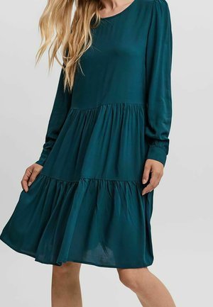 Femme portant une robe longue à manches longues de couleur turquoise, à volants et longueur genou, avec un col rond, posant devant un fond clair uni.