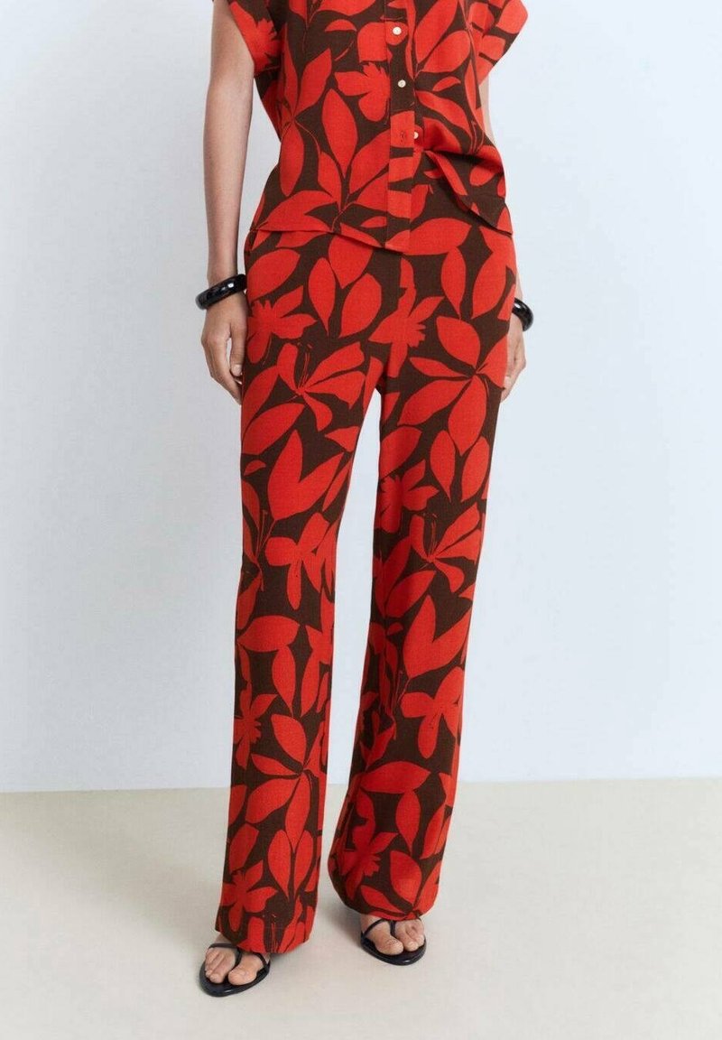 Femme portant un pantalon large à motifs floraux rouges et brun foncé avec une chemise boutonnée à manches courtes assortie et des sandales noires.