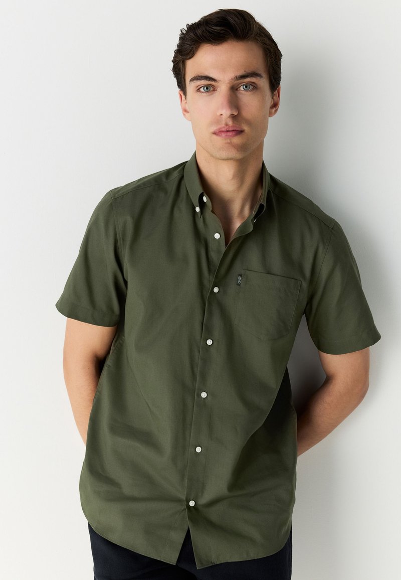 Chemise à manches courtes, vert olive, en coton. Comprend un col boutonné, une poche poitrine et des boutons blancs sur la patte de boutonnage avant.