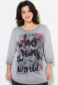 Chemise à manches longues grise avec un texte graphique audacieux en noir et rouge, "qui dirige le monde", sur un fond texturé et à motifs.