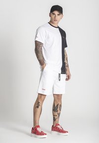 Čierno-biela farbne blokovaná tričko, biele šortky s logom, červené tenisky s popisom a čierna čiapka, prezentujúce tetované rámy.