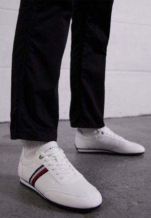 Pies con zapatillas blancas con rayas rojas y azul marino, combinadas con calcetines blancos y pantalones negros, de pie sobre un suelo de concreto gris.