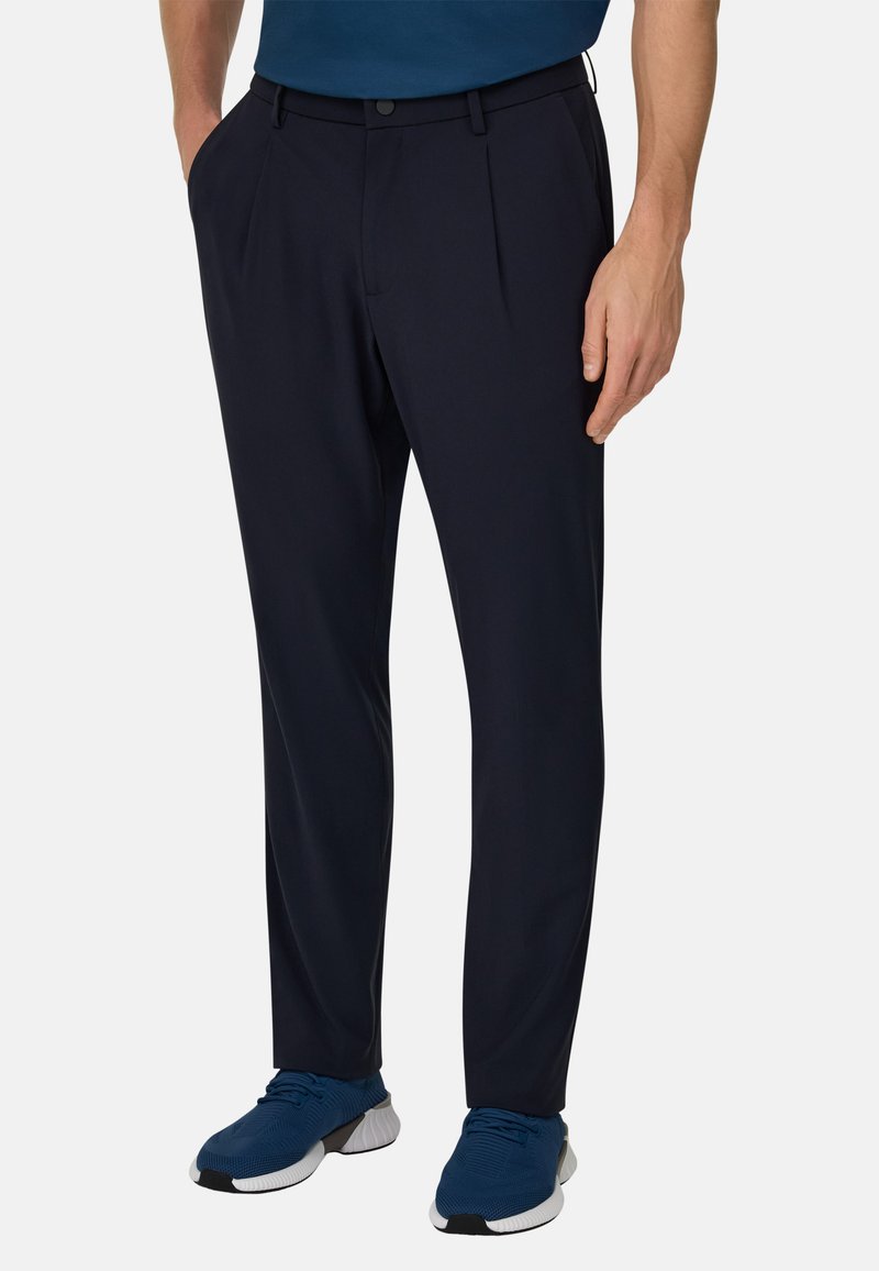 Pantalon bleu marine en tissu léger, avec plis, poches latérales et coupe ajustée, associé à des chaussures de sport bleues.