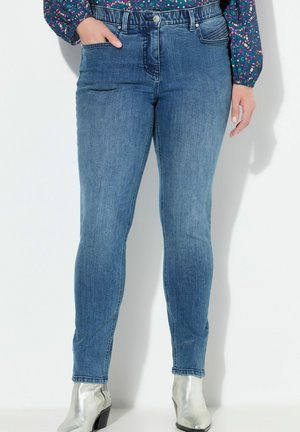 Nainen, joka käyttää sinisiä kapeita farkkuja etutaskuilla, värikästä kukkakuosista puseroa ja hopeisia nilkkureita, seisoo valkoista taustaa vasten.