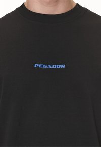 Černé bavlněné tričko s modrým logem "PEGADOR" na hrudi. Krátké rukávy a žebrovaný kulatý výstřih. Hladký povrch.
