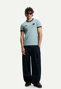 Light blue fitted t-shirt with navy ringer collar and logo, paired with loose dark denim trousers and black shoes, model wearing sunglasses.