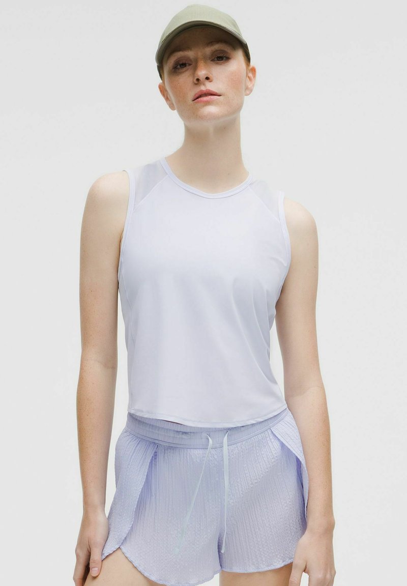 Witte mouwloze sporttop en lichtpaarse gewatteerde shorts. Model draagt een lichtgroene pet. Beide stukken hebben een comfortabele pasvorm.