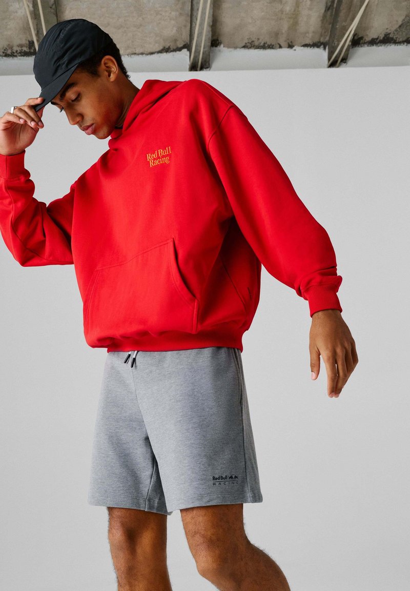 Κόκκινο φλις hoodie σε φωτεινό κόκκινο χρώμα με τσέπη κάγκουρου και χρυσό λογότυπο "Red Bull Racing". Συνδυάζεται με γκρι βαμβακερά σορτς που φέρουν λογότυπο.
