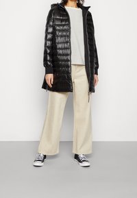 Czarna puchowa kurtka o pikowanej fakturze, z kapturem i kieszeniami na zamek, noszona na jasnoszarym, prążkowanym topie oraz beżowych spodniach o szerokich nogawkach, z czarnymi trampkami.