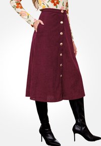 Mujer vistiendo una falda midi burdeos abotonada, una blusa de manga larga con estampado floral y botas de rodilla negras de tacón alto, con la mano en el bolsillo.