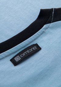 Světle modrý bavlněný límec trička s černým žebrovaným lemováním. Obsahuje černou látkovou štítek s nápisem "ombre" v šedém textu. Hladká textura.