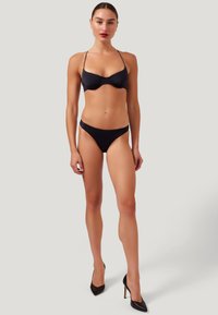 Frau in schwarzem Bikini und schwarzen High Heels vor einem schlichten hellgrauen Hintergrund, die frontal steht und die Arme entspannt hält.