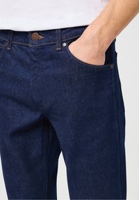 Tmavě modré džíny z denimu s klasickým designem, vybavené předním kapsou, viditelným šitím a kovovým knoflíkem v pase.